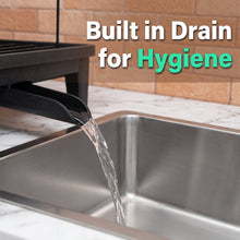 A close-up of a kitchen sink with water flowing from a black built-in drain spout into the sink. A dark dish rack sits above, and the background shows a tan tile wall and white marble countertop.
